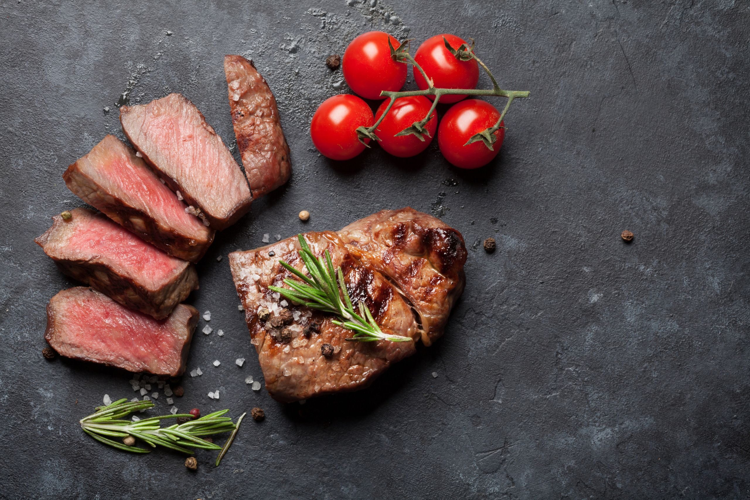 Grilled steak sliced into portions, accompanied by cherry tomatoes and fresh rosemary on a dark stone surface. A sprinkle of coarse salt and pepper can also be seen.