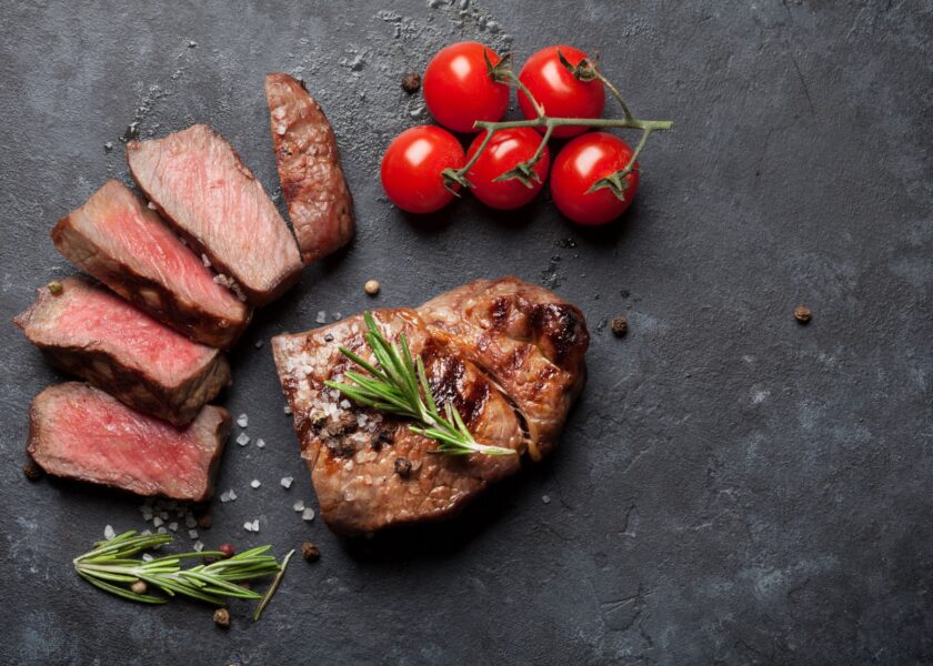 Grilled steak sliced into portions, accompanied by cherry tomatoes and fresh rosemary on a dark stone surface. A sprinkle of coarse salt and pepper can also be seen.