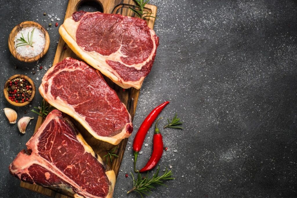 What Are the Different Types of Steaks? 1 - Driven A wooden cutting board featuring three cuts of raw beef steak, along with red chilies, garlic cloves, and bowls of salt and mixed peppercorns, set against a dark surface.