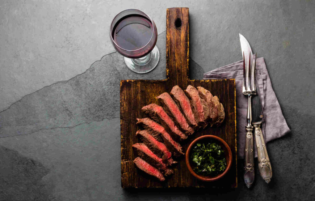 Best Wines to Pair with Ribeye, Filet, and Sirloin 1 - Driven Sliced steak arranged on a wooden cutting board next to a bowl of green sauce, accompanied by a glass of red wine and silver utensils placed on a gray napkin.