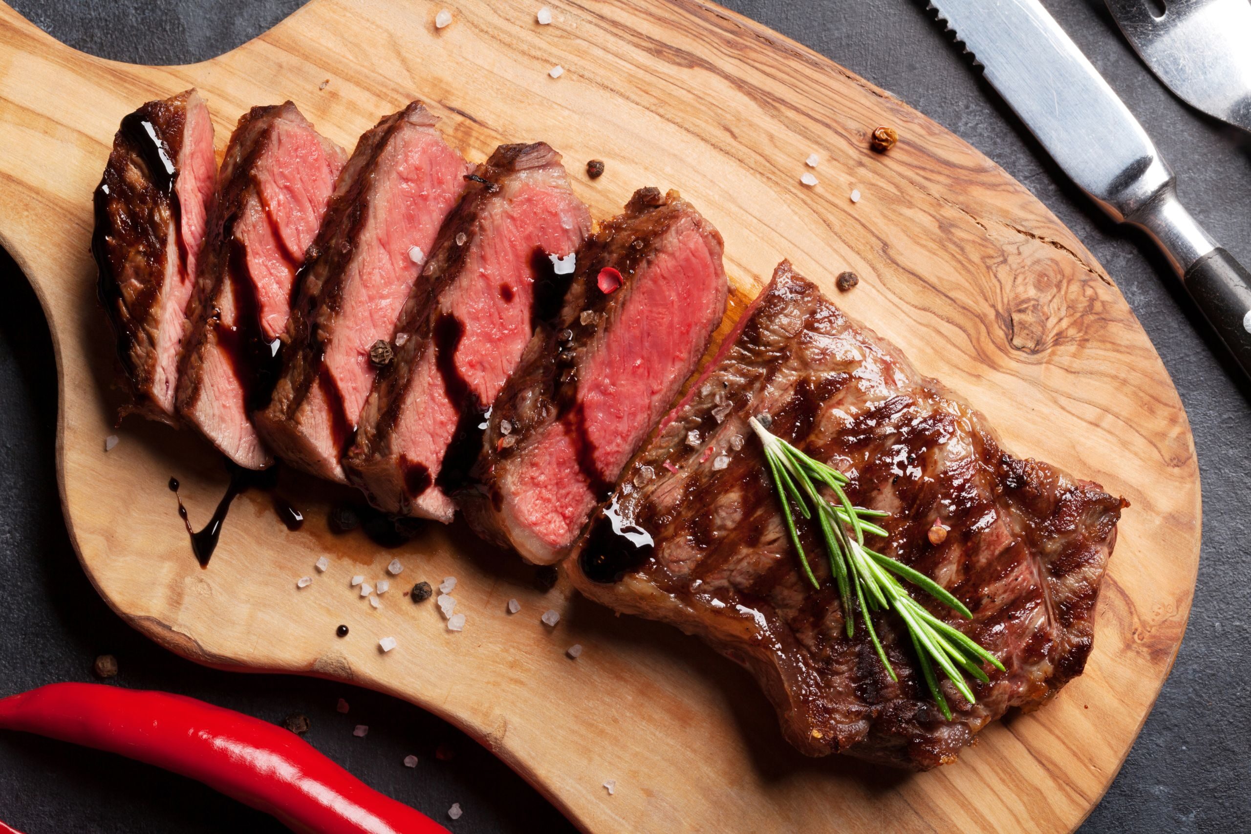 Sliced steak arranged on a wooden cutting board, featuring pink interior and grill marks. A sprig of rosemary is placed on the steak, with seasoning and red chili peppers scattered around.