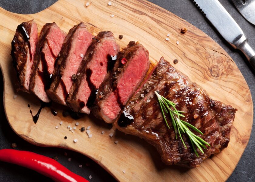 Sliced steak arranged on a wooden cutting board, featuring pink interior and grill marks. A sprig of rosemary is placed on the steak, with seasoning and red chili peppers scattered around.