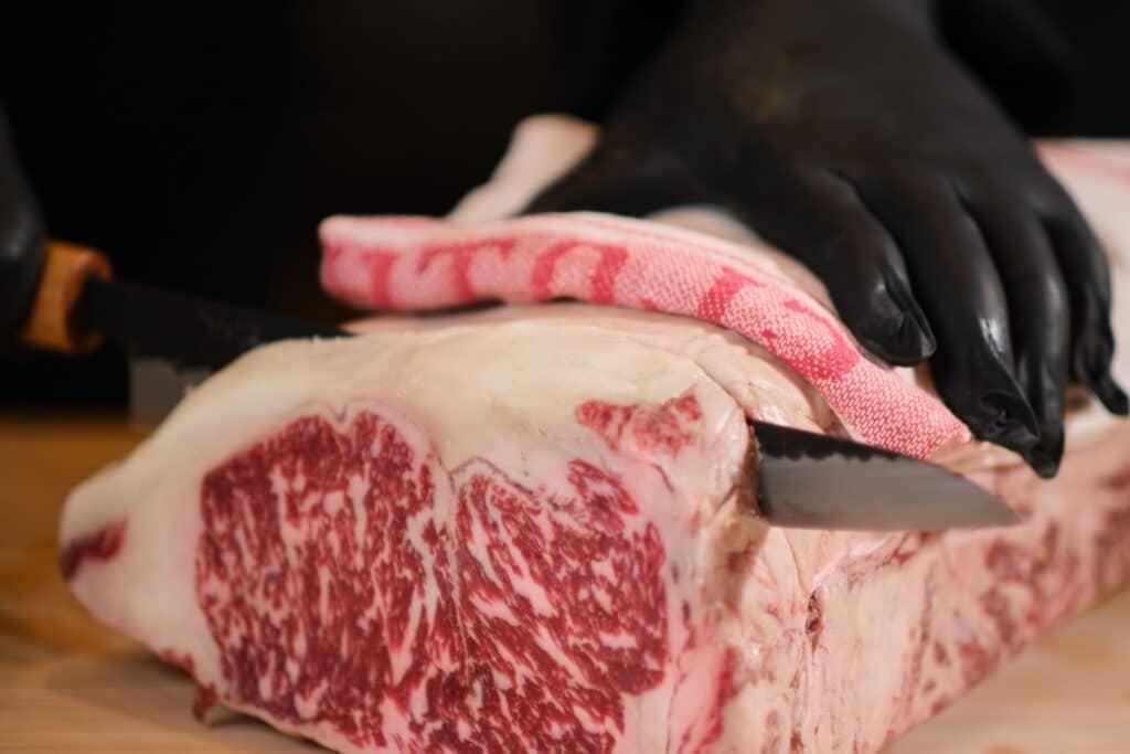 Wagyu Beef: What is it and Where Does it Come From? 1 - Driven A close-up of a marbled piece of meat being sliced with two knives, while a person wearing black gloves holds the meat steady. A red and white checkered cloth is seen on top of the meat.