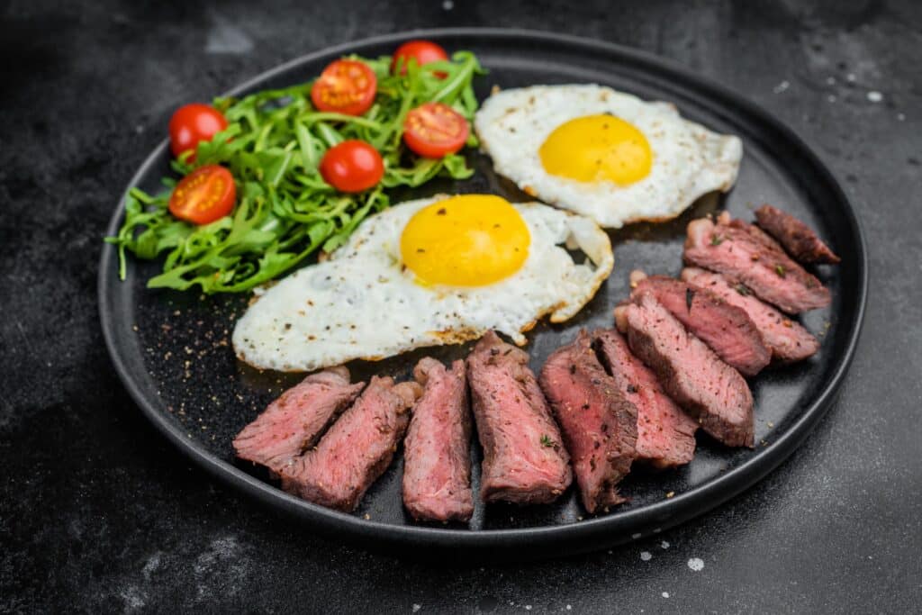 Endless Brunch: Spend Sunday Morning at Driven 1 - Driven A black plate with two fried eggs, cherry tomatoes, and slices of cooked steak arranged beside a bed of arugula.