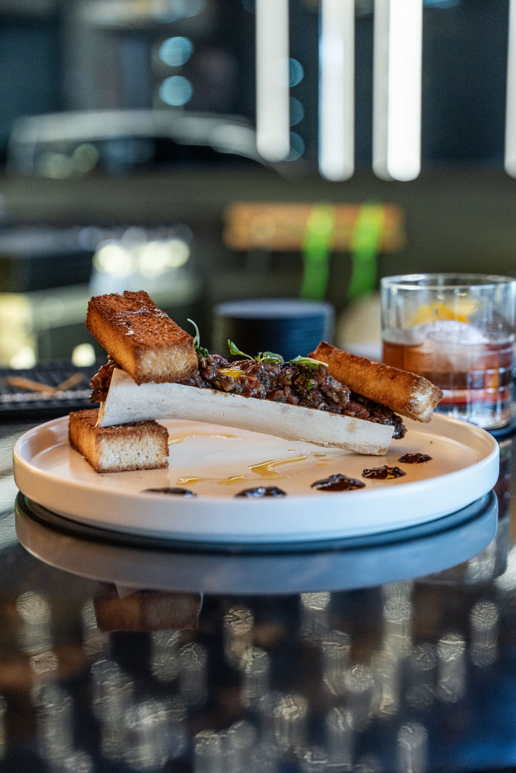 A modern plate featuring a piece of toasted bread alongside a portion of cooked beef and garnishes. The dish is presented on a white plate with a drizzle of sauce and appears on a reflective black surface. A glass with a yellow drink is visible in the background.