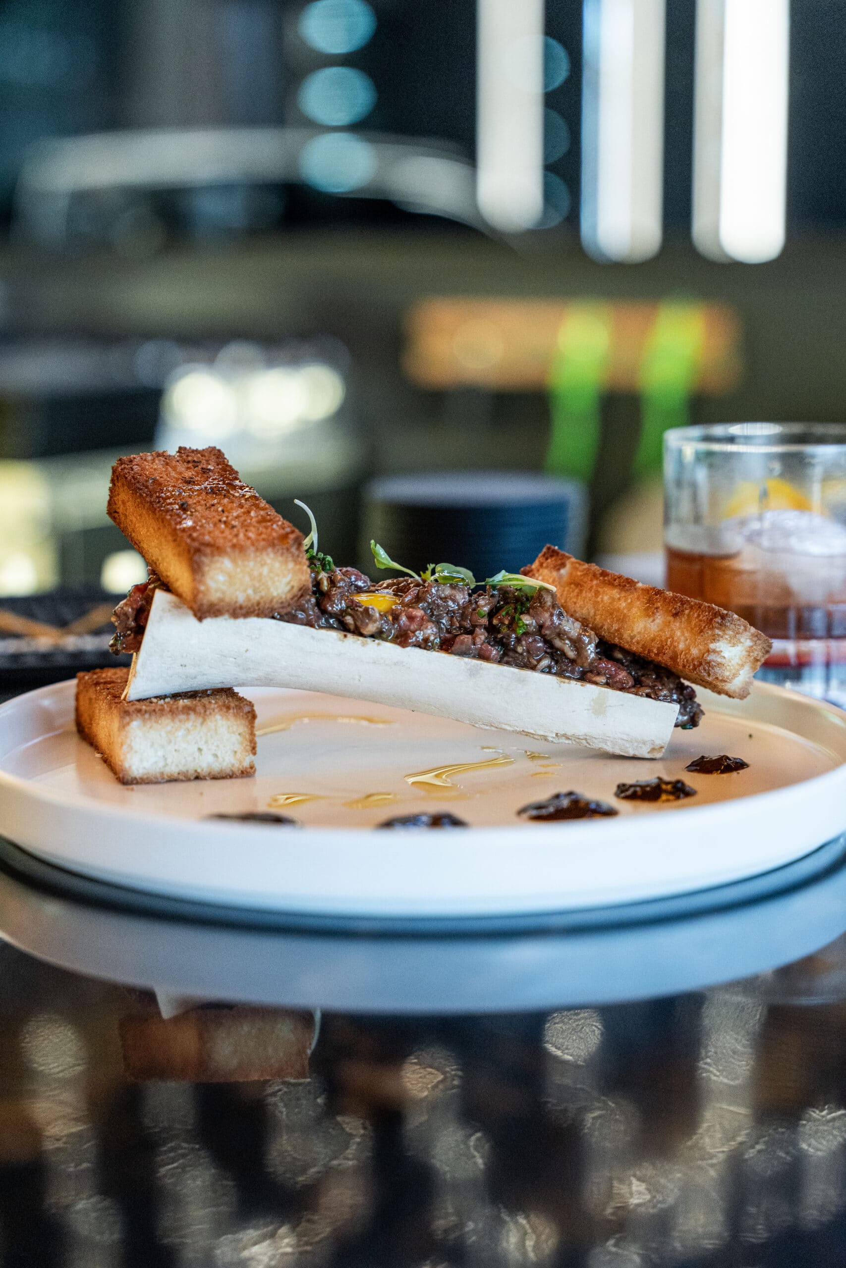 A plate featuring a large piece of bone marrow with a crusty piece of toasted bread on one end. The bone is filled with a mixture of ground meat and herbs. Dark sauce and small servings of additional ingredients are artfully arranged around the plate.