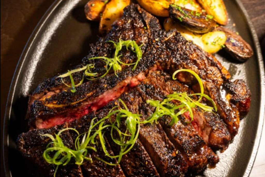 History of Steakhouse Dining 1 - Driven A sliced grilled steak topped with green garnish on a dark plate, with roasted potatoes and mushrooms in the background.