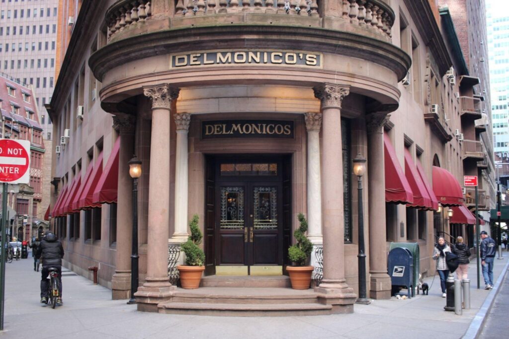 History of Steakhouse Dining 2 - Driven The entrance to Delmonico’s restaurant with columns, red awnings, and potted plants on a city street.