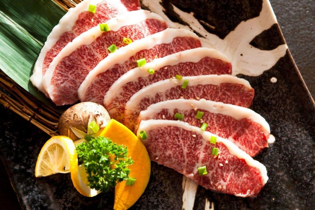 History of Steakhouse Dining 3 - Driven Sliced marbled beef arranged on a black plate with a lemon wedge, parsley garnish, and a mushroom beside a green leaf.